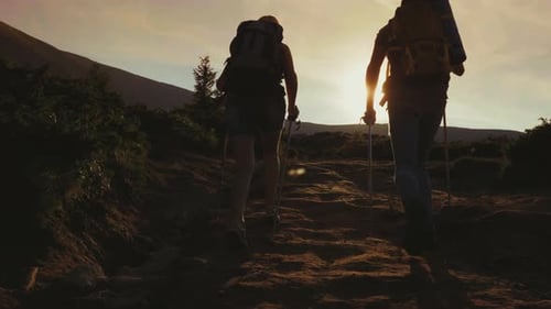 Two Travelers with Backpacks Go Forward Along the Mountain Path Towards the Sun