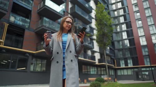 Formally Dressed Woman Walking Down the Street in a Business District with Coffee in Hand and Using