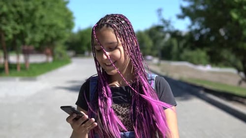 A Beautiful Girl with Long Pigtails Walks in the Park Using a Mobile Phone