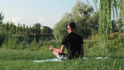 Back view of young man sitting on yoga mat in lotus position and meditating in city park.