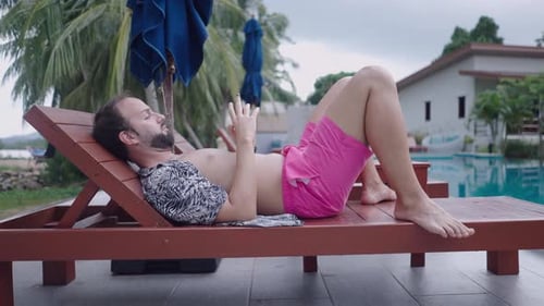Bearded Man Traveler Watching Video on Phone While Resting on Wooden Deckchair By the Pool in
