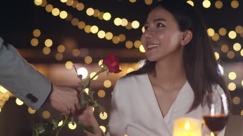 Young asian couple surprise moment with rose flower at romantic dinner in rooftop restaurant