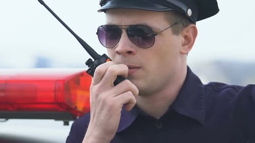 Police Officer Talking on Walkie-Talkie Near Police Car