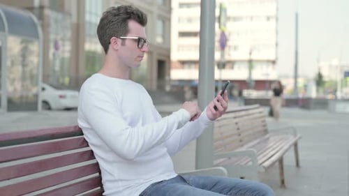 Side View of Young Man Using Smartphone While Sitting Outdoor on Bench