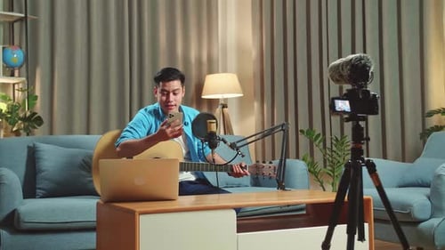 Un hombre asiático es un vlogger con guitarra Lee un comentario en un teléfono móvil mientras transmite en vivo