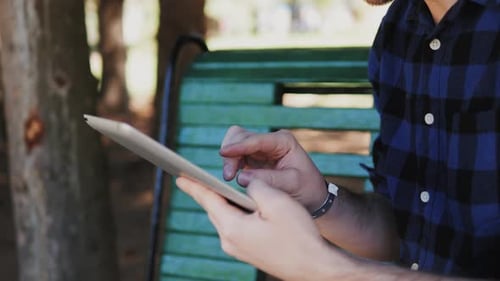 Young Man Sitting on the Bench Near Grass in Park with Tablet