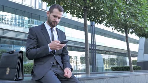 Businessman Sits with Mobile Phone Near Office Building