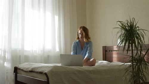 Freiberuflerin sitzt auf dem Bett und benutzt Laptop. Eine Frau arbeitet oder studiert remote.