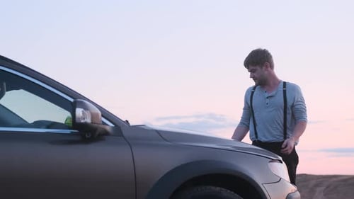 Bearded Country Man in Gray Turtleneck or Shirt and Black Suspenders Walks Up to Hood of Car at Dusk