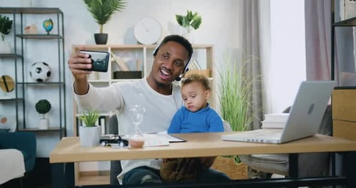 Padre de piel negra con auriculares haciendo una selfie junto con su hijo pequeño en casa