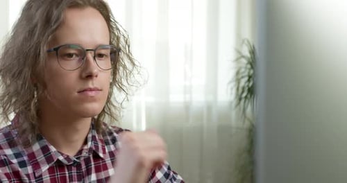 A pensive young man in glasses looks at the computer screen. Face of a student, close-up