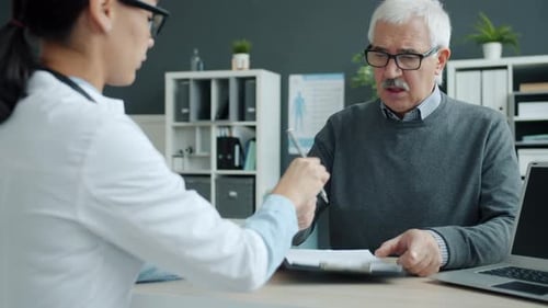 Senior Man Patient Signing Medical Document Talking To Female Doctor in Clinic