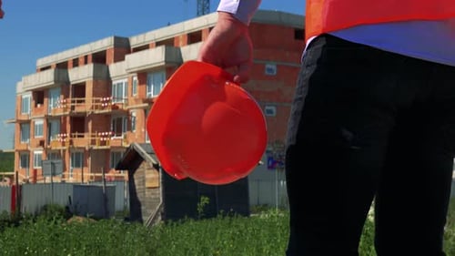 Construction Worker Holds Hardhat in Hand and Stands in Front of Building Site