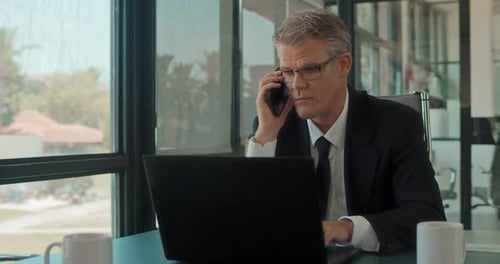 A businessman talking on the phone while working on a laptop at the office
