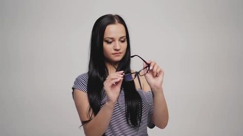 brunette in striped dress is putting on eyeglasses on a gray background