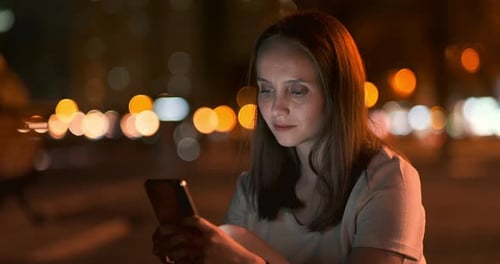 A Young Girl on the Background of the Night City Looks Into the Smartphone Screen