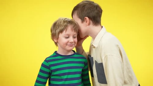 Niños sonrientes y positivos que comparten secretos susurrando al oído sobre fondo amarillo