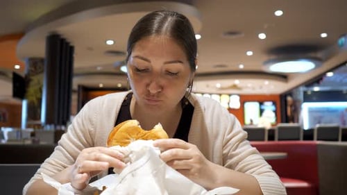 Young Brunette Woman is Eating Burger in Fast Food Restaurant in Food Court