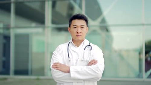 Close up portrait of Asian doctor looking at camera with arms crossed on background