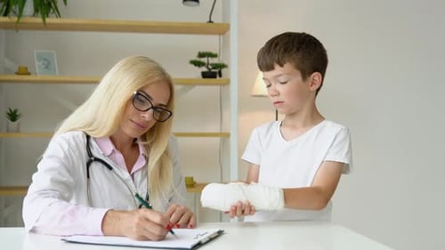 Smiling Senior Female Family Doctor in Uniform Consulting Little Kid Patient with a Broken Hand at