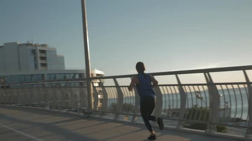 Happy attractive young woman in blue sportswear is running on bridge in the morning at sunrise.
