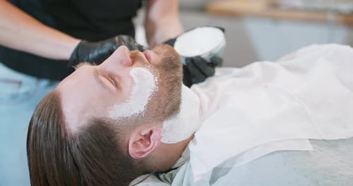 Nahaufnahme der Hände einer Friseurin, die mit Rasierschaum auf das Gesicht eines jungen bärtigen Mannes aufträgt und