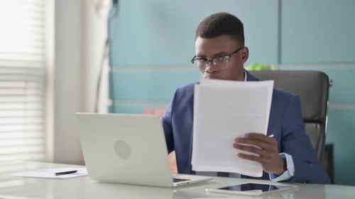Young African Businessman with Laptop Reading Documents in Office