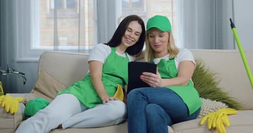 Dos mujeres limpiadoras descansan en el sofá de una casa contemporánea y ven videos divertidos en una tableta
