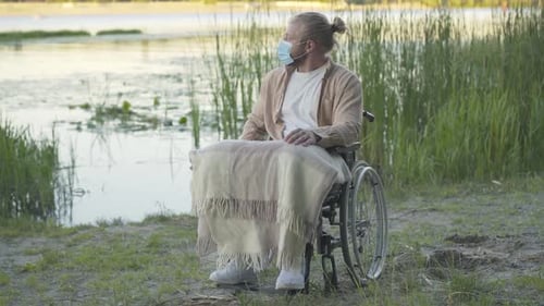 Wide Shot Portrait of Paralyzed Man on Wheelchair in Face Mask Resting on River Bank at Sunset