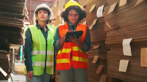 Two female workers in safety uniforms check a pile of stock in a paper factory.
