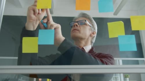 Mature Businessman Writing on Sticky Notes on Office Glass Wall Busy with Business Planning