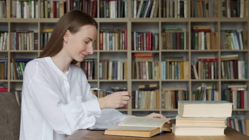 Young Woman Sitting at Table with Books and Laptop in University Library and Studying