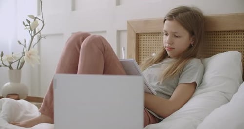 Müde und gelangweilte Schülerin liegt auf dem Bett mit Heftbuch und Hörstunde auf Laptop, e.