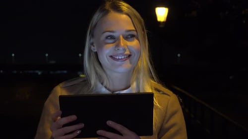 A Young Beautiful Woman Works on a Tablet in an Urban Area at Night, Looks Up and Smiles at Camera