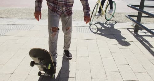 Two diverse male friends riding a skateboard and bicycle in the street