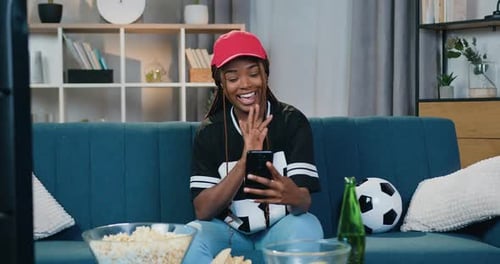 Mujer afroamericana en ropa deportiva sentada en el sofá de casa y hablando por teléfono