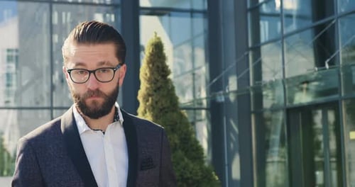 Empresario con gafas que posa ante la cámara cerca de un moderno edificio de cristal durante una pausa para tomar un café