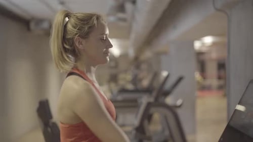 Sportive Young Caucasian Woman Drinking Water As Walking on Treadmill in Gym. Side View of Beautiful