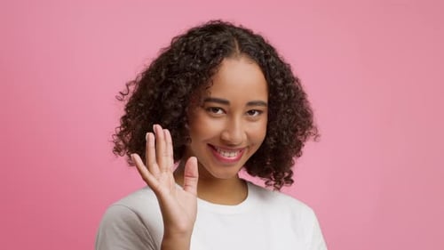 Verspielte Afroamerikanerin winkt Hallo lächelnd auf rosafarbenem Hintergrund