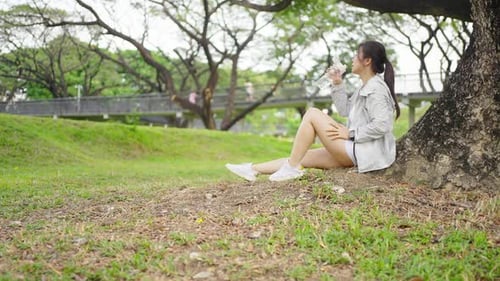 4K Asian woman drinking water from a bottle while jogging at public park in the morning.