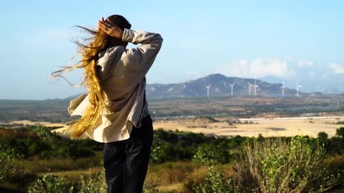 Woman with long red hairs on hilltop watching eco friendly wind turbine farm in background - Windy d