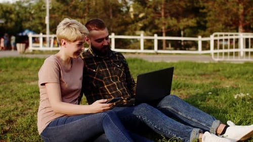Hipster Man and Caucasian Woman are Sitting on Lawn in Park Talking Smiling and Using Laptop