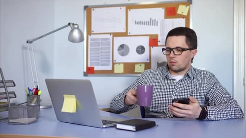 Office Worker Sitting and Using Smartphone. He Take a Break and Drink Coffee.