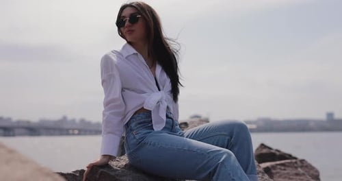 Stylish Woman Sitting on Rocky Shoreline by City