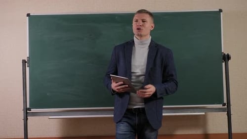 A Young Male Teacher Stands in the Lecture Room with a Tablet in His Hands