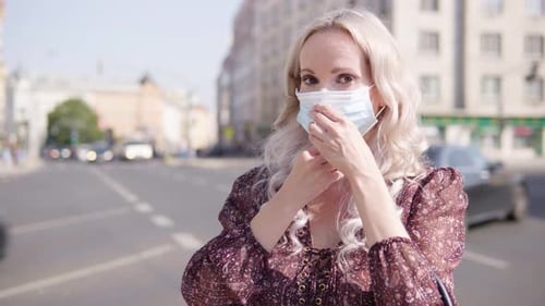 A Middleaged Caucasian Woman Puts on a Face Mask and Looks at the Camera in an Urban Area