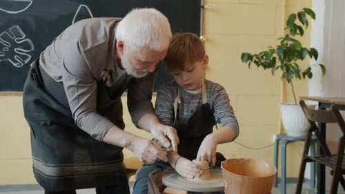 Experienced Potter Teaching Small Boy To Make Ceramic Vase on Throwing Wheel in Studio