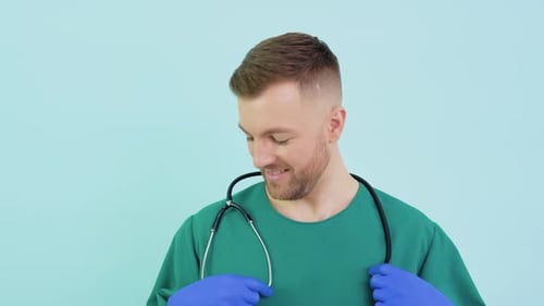 Erfolgreicher Doktorchirurg in grüner Uniform, blauen Handschuhen und Stethoskop schaut in die Kamera und lächelt weiter