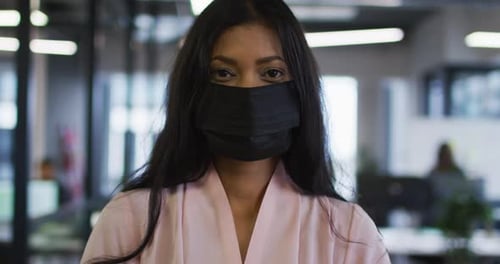 Portrait of mixed race businesswoman wearing face mask standing in office looking to camera