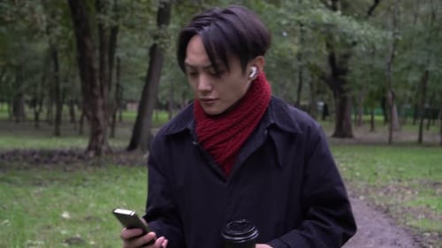 Attractive Asian Man in a Coat Uses a Smartphone in an Autumn Park